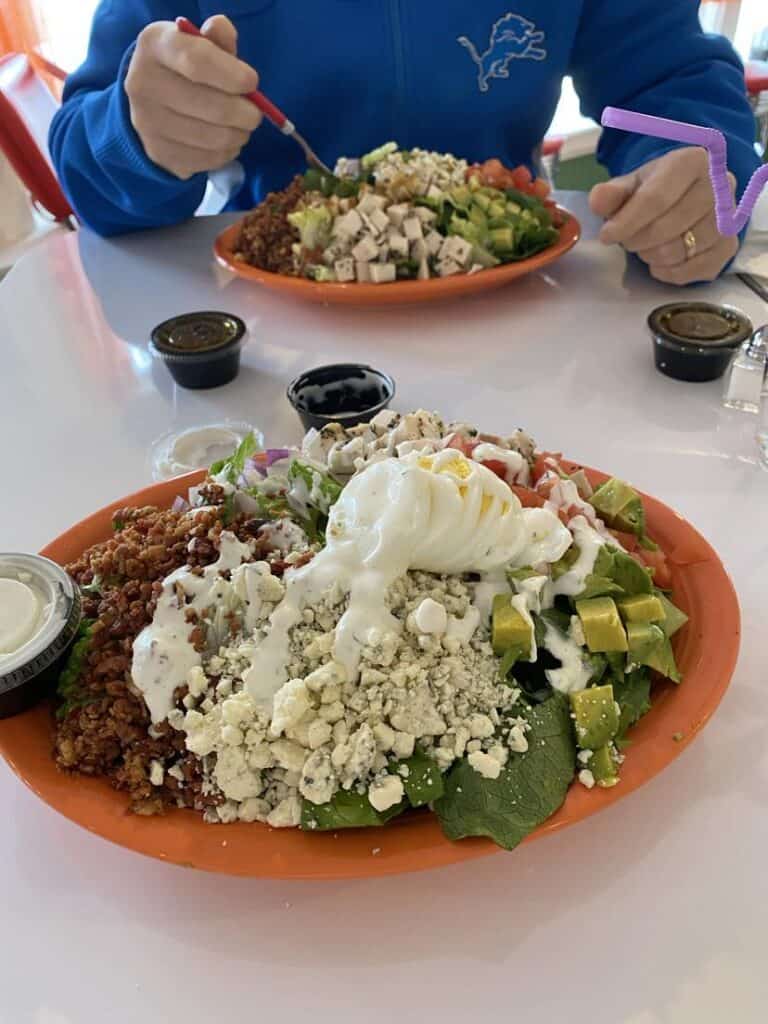 Two plates of salad on a table, topped with quinoa, avocado, cheese, and dressing—some of my favorite things. A person in a blue jacket is holding a fork, and dressing cups are nearby.