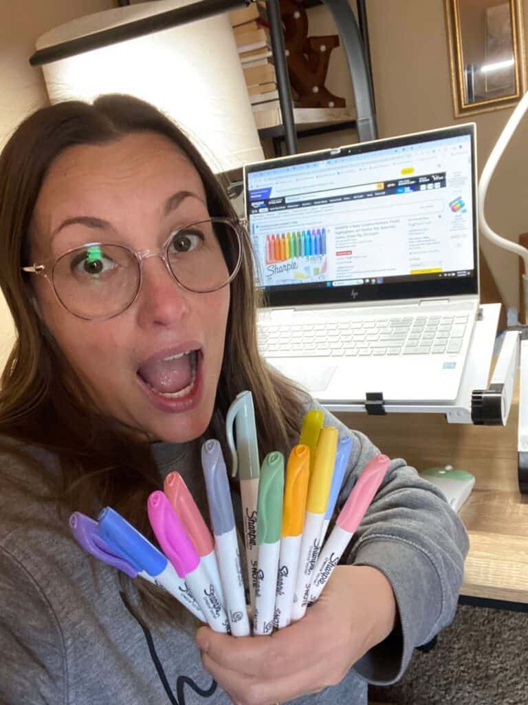 A person with glasses holds a variety of colorful markers, expressing excitement over a recent post. In the background, a laptop displaying a website is visible on the desk, suggesting the vibrant creativity they bring to each personal update.
