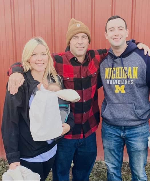 Three adults and a baby in front of a red wall. One person is holding the baby, another in a beanie and plaid shirt, and the third wearing a Michigan Wolverines hoodie.