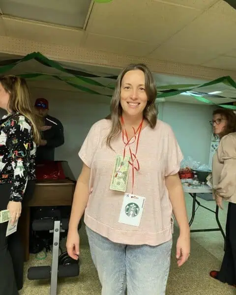 A woman stands indoors wearing a red lanyard adorned with money and a Starbucks card picture, symbolizing a personal journey. In the background, people mingle around tables strewn with various items, creating an atmosphere that warms the heart.