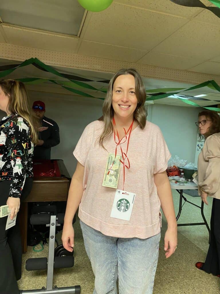 A woman stands indoors wearing a red lanyard adorned with money and a Starbucks card picture, symbolizing a personal journey. In the background, people mingle around tables strewn with various items, creating an atmosphere that warms the heart.