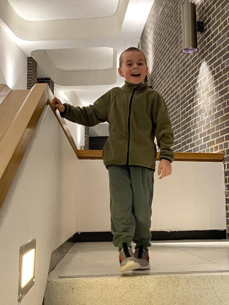 A smiling child in a green outfit stands on a stairway indoors, holding the railing with one hand, as if ready for Liam Takes the Stage. The walls are a charming mix of white and brick, creating a cozy backdrop for this favorite moment.