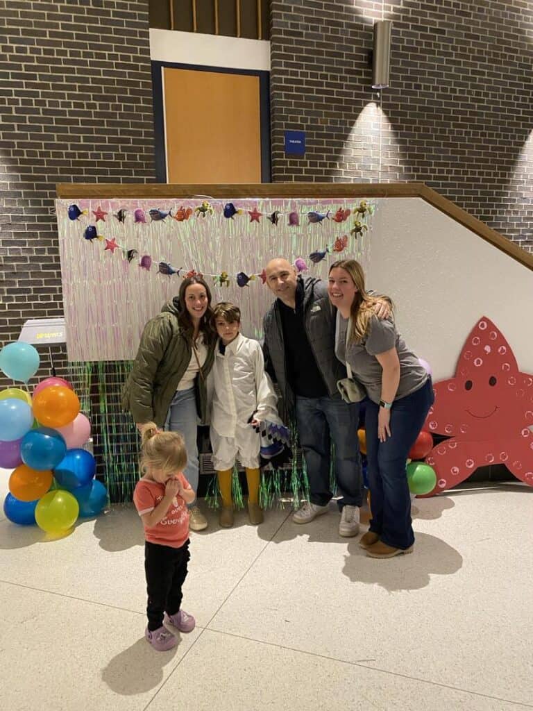 A group of four adults and one child pose in front of a festive backdrop with balloons and a large red star decoration, reminiscent of some of my favorite things.
