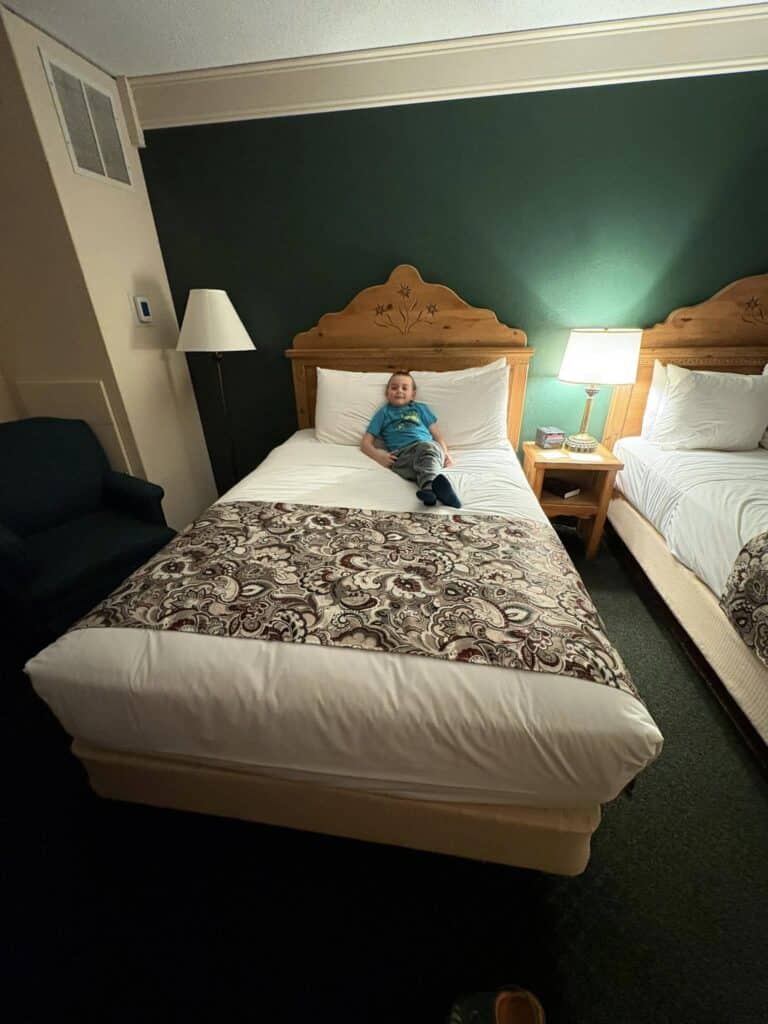 A child in a blue shirt sits on a neatly made bed in the dimly lit room of the Bavarian Inn, surrounded by green walls. The cozy room features two beds and a softly glowing bedside lamp, creating a serene ambiance.