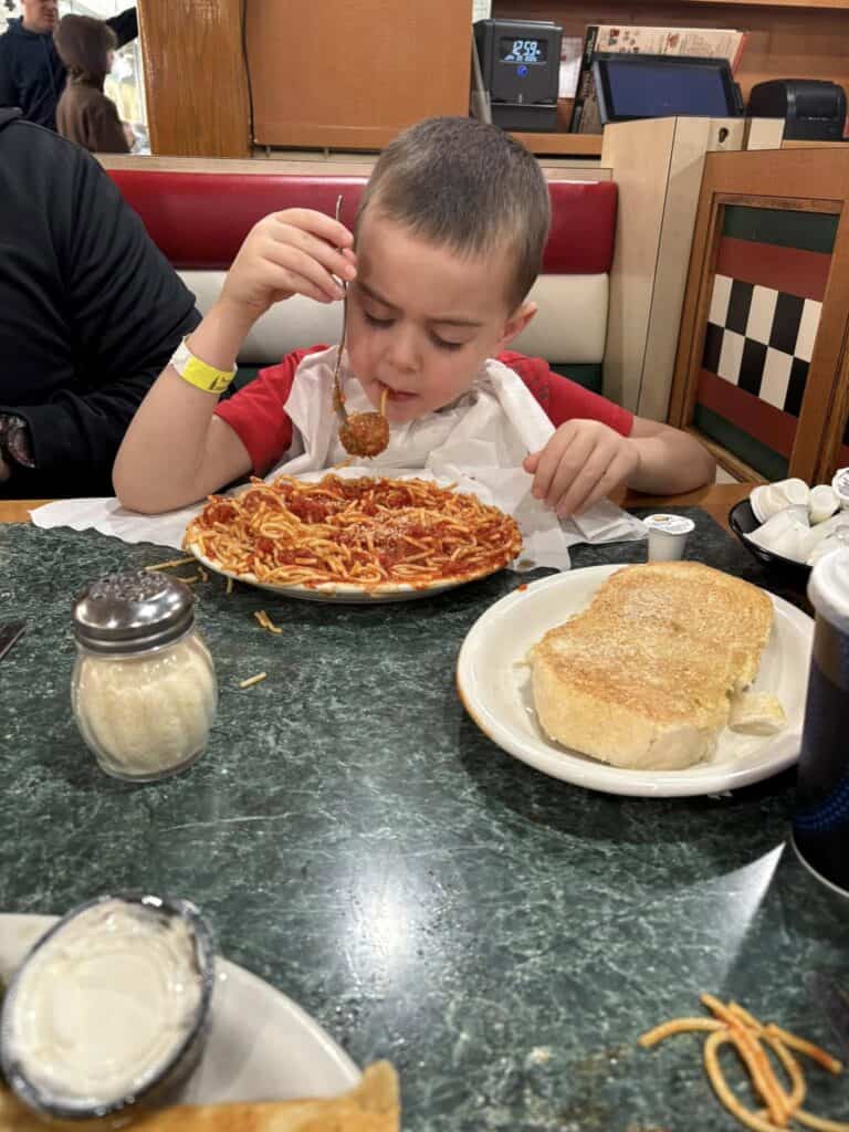 A young boy in a red shirt enjoys spaghetti with a meatball at the Bavarian Inn's cozy diner table, with bread and a drink nearby.