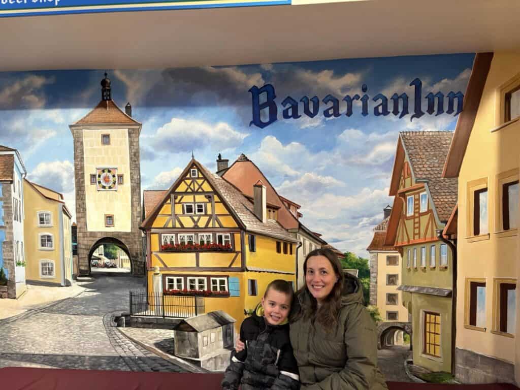A woman and a child sit in front of a mural depicting a charming Bavarian Inn nestled within a picturesque village scene, complete with timber-framed houses and a towering structure.