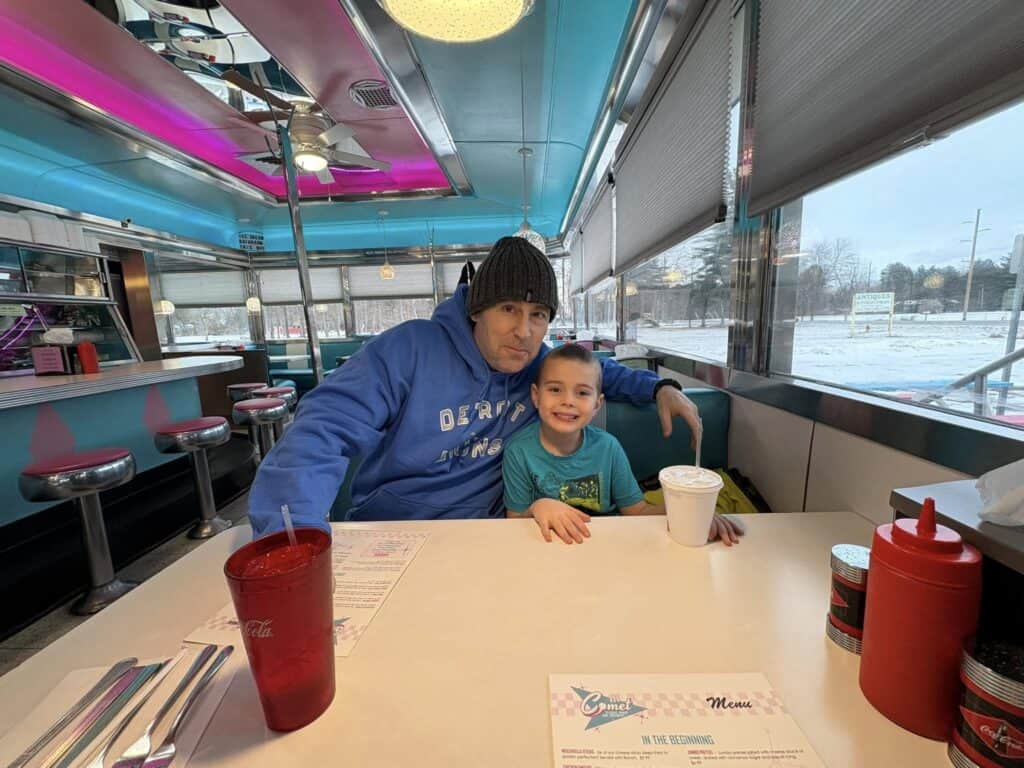 A man and a child sit smiling at a diner booth in the cozy Bavarian Inn. A menu and drinks rest on the table amid retro-style decor with pink and teal accents. Snow gently blankets the world outside the window, adding to the charming ambiance.