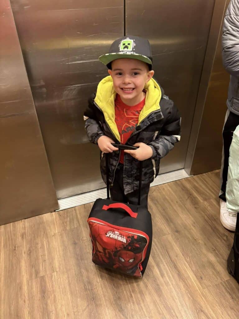 A child in a hat stands in an elevator at the Bavarian Inn, clutching a Spider-Man themed suitcase. The child beams with joy, wearing a jacket over a red shirt.