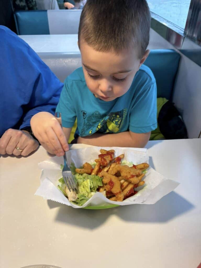 A young boy in a blue shirt enjoys a salad and fries with a fork at a Bavarian Inn Restaurant booth.
