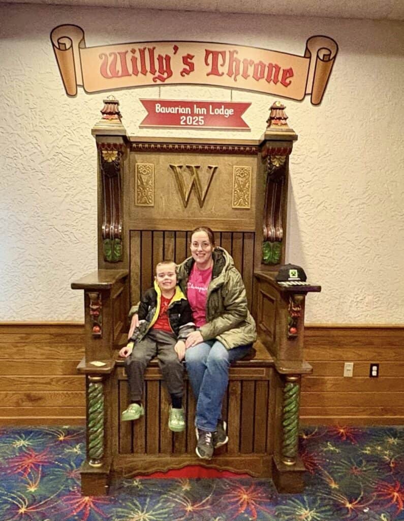 Two people sit on a large wooden throne labeled "Willy's Throne" at the Bavarian Inn Lodge. This majestic seat is ornately decorated, with a "W" emblem on the backrest and a charming hat casually resting on the armrest, perfectly capturing the enchanting spirit of the Bavarian Inn.