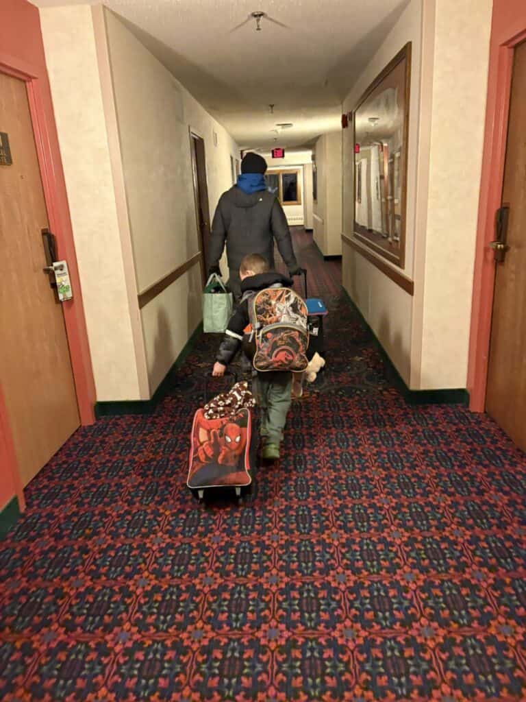 Two people are walking down the hotel hallway of the Bavarian Inn, each carrying a suitcase and wearing backpacks. The corridor boasts a patterned carpet and framed pictures adorning its walls, creating a warm and inviting atmosphere.