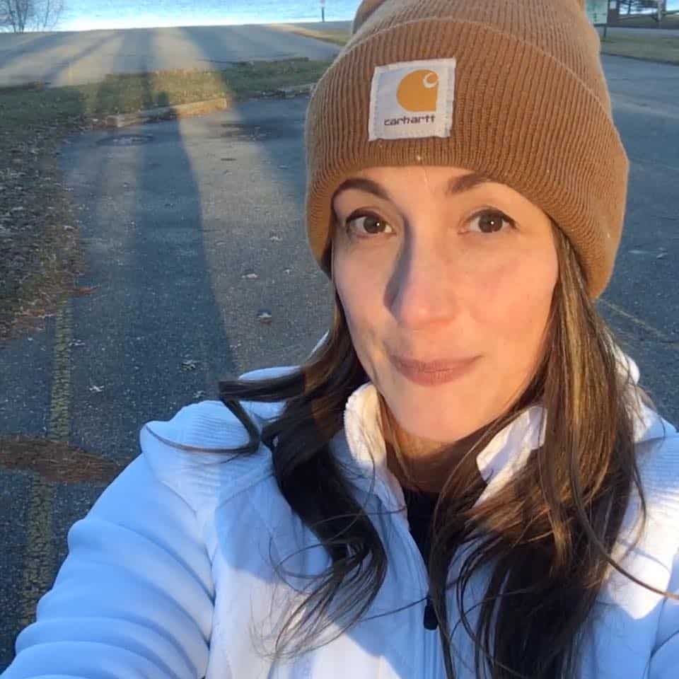 A person wearing a brown beanie and white jacket snaps a sunny selfie on a paved path, capturing the serene backdrop of grass and trees—an ideal life update from an otherwise ordinary day.