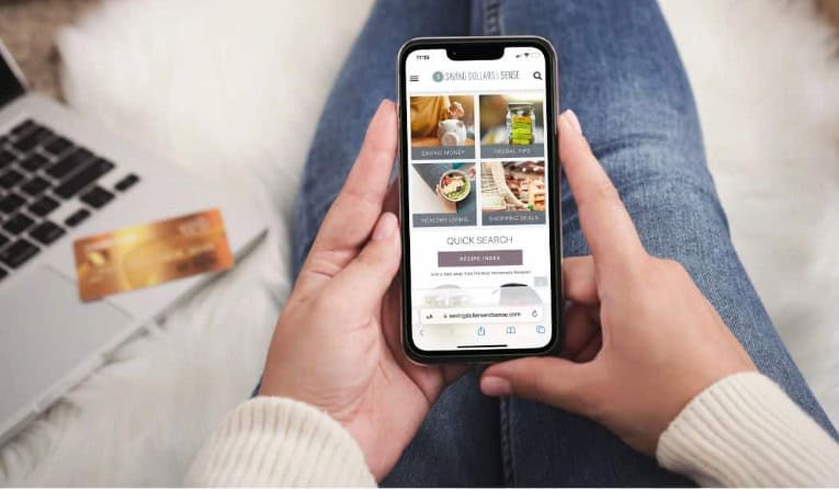 A person is holding a smartphone displaying a recipe website, embarking on a culinary journey, with a laptop and credit card nearby.