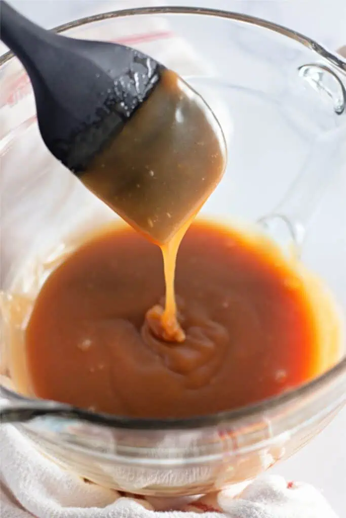 A silicone brush drips rich, homemade caramel sauce into a clear glass bowl on a white surface, turning every moment into an easy caramel recipe masterpiece.