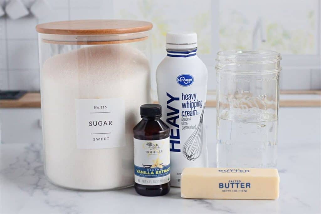Ingredients for a homemade caramel recipe on a kitchen counter: a jar of sugar, bottle of heavy whipping cream, vanilla extract, glass of water, and a stick of salted butter.