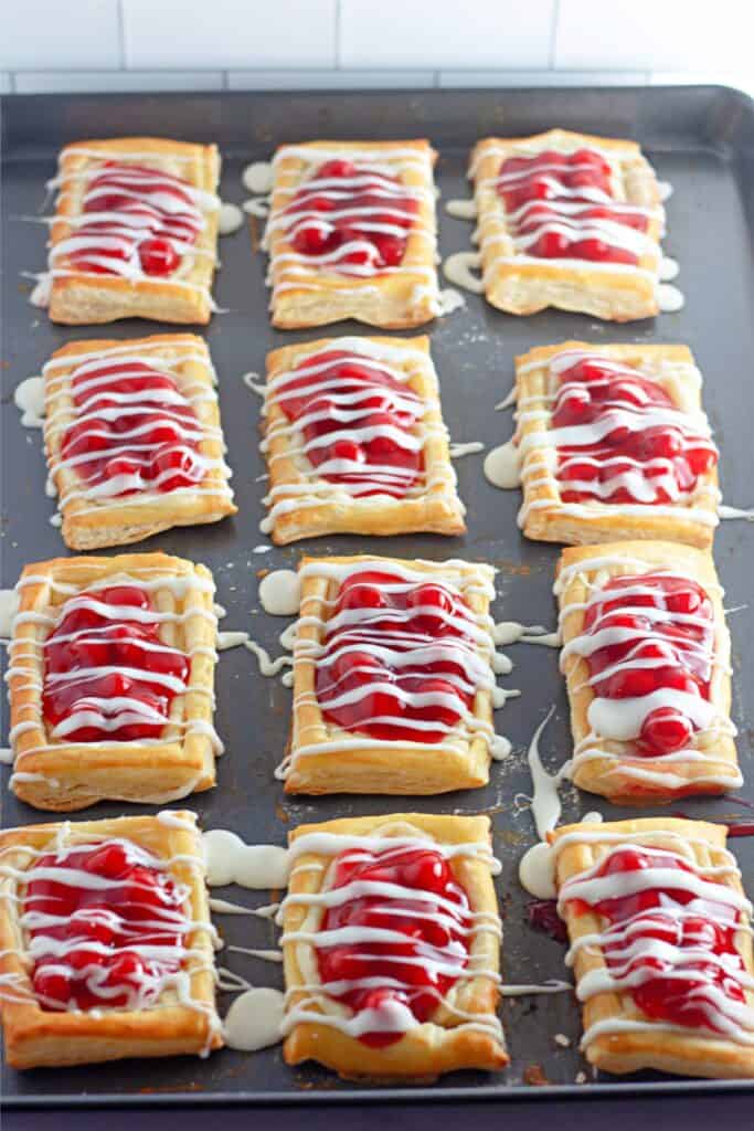 Twelve Cherry Cheesecake Danish pastries with white icing drizzled on top, arranged in rows on a baking sheet, offering a delicious twist to the classic recipe.