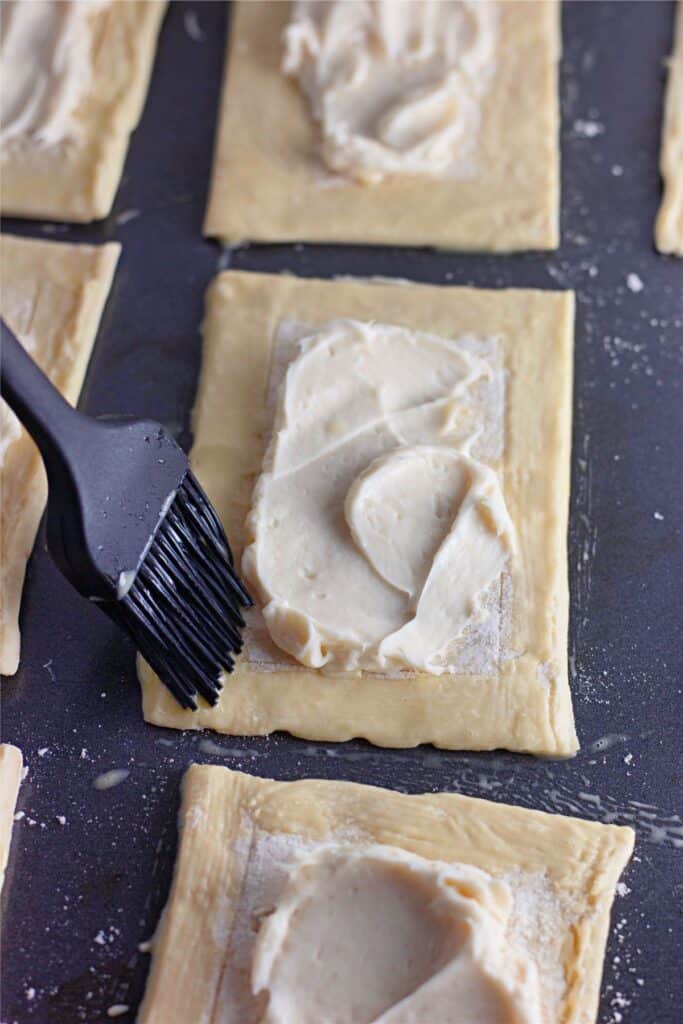 Square pieces of dough topped with a delightful cream cheese mixture are gently brushed with a sleek black pastry brush, setting the stage for a mouthwatering Cherry Cheesecake Danish recipe.
