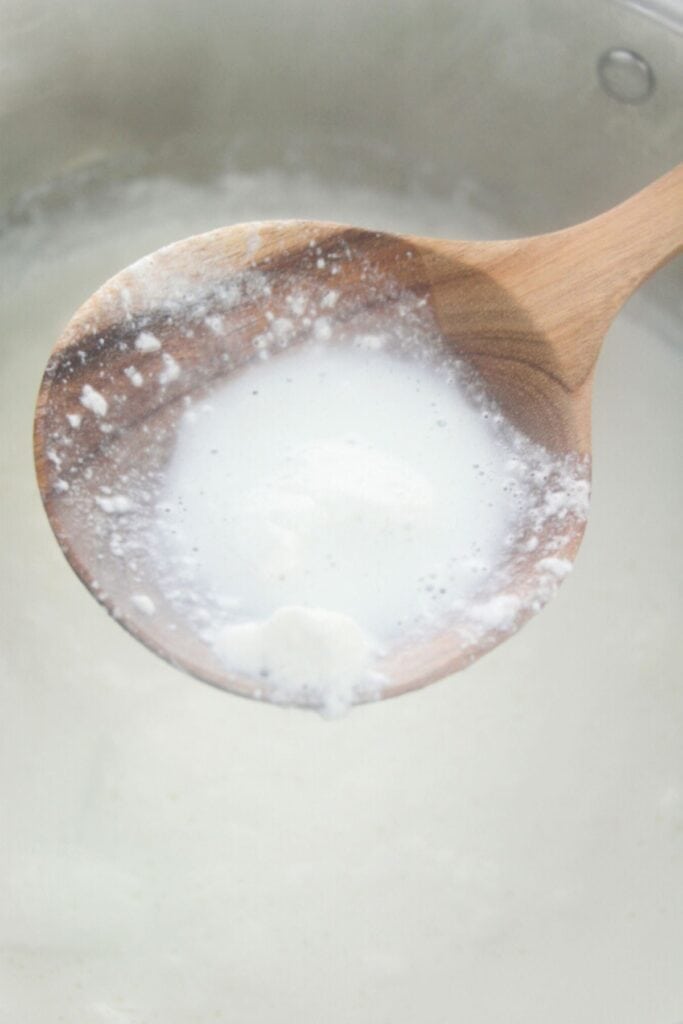 Close-up of a wooden spoon scooping a thick, white homemade ricotta from a pot, resembling yogurt or cream.