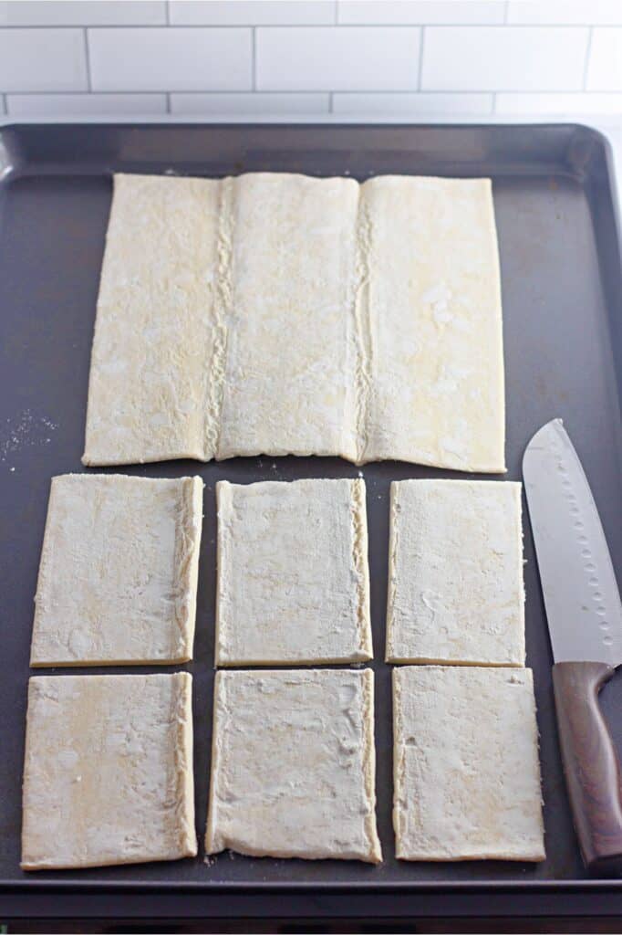 Dough placed on a baking sheet is partially cut into rectangles, ready to transform into delightful Cherry Cheesecake Danishes. A knife lies next to it, awaiting the final touch.