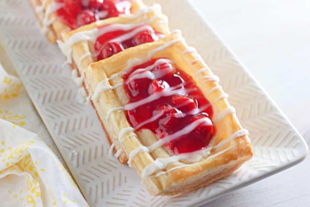 Cherry cheese Danish pastries grace a white patterned platter, each treat topped with a delicate white icing drizzle.