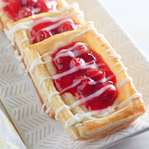 Cherry cheese Danish pastries grace a white patterned platter, each treat topped with a delicate white icing drizzle.