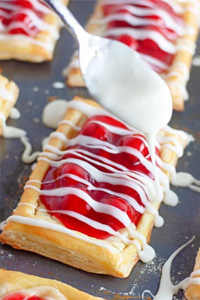 Cherry pastries with icing being drizzled over them from a spoon rest on a dark surface, resembling a Cherry Cheesecake Danish straight out of an exquisite recipe.