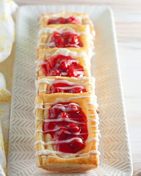 Four Danish pastries topped with cherry cheesecake filling and drizzled with white icing are arranged in a row on a rectangular plate.