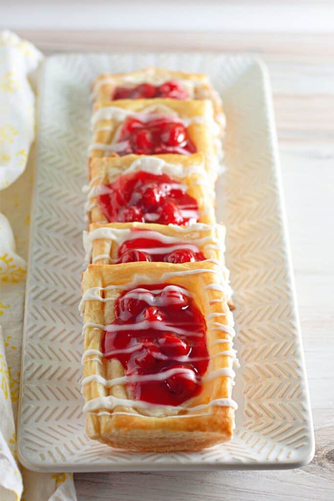 Four Danish pastries topped with cherry cheesecake filling and drizzled with white icing are arranged in a row on a rectangular plate.