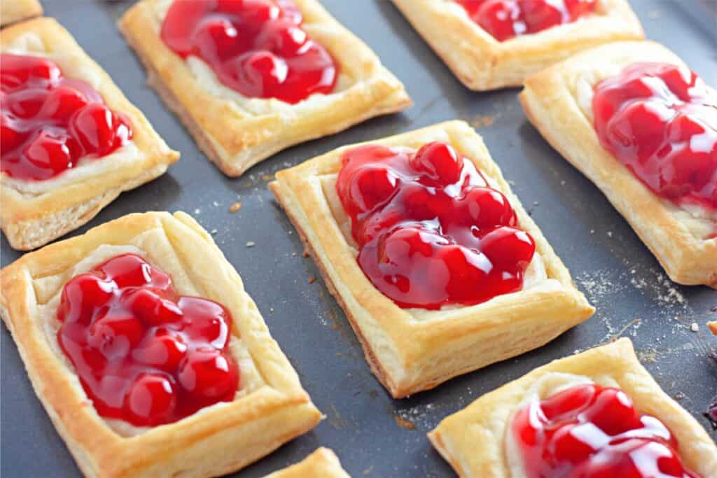 Rectangular pastries topped with bright red cherry filling, reminiscent of a Cherry Cheesecake Danish, laid out on a baking sheet.