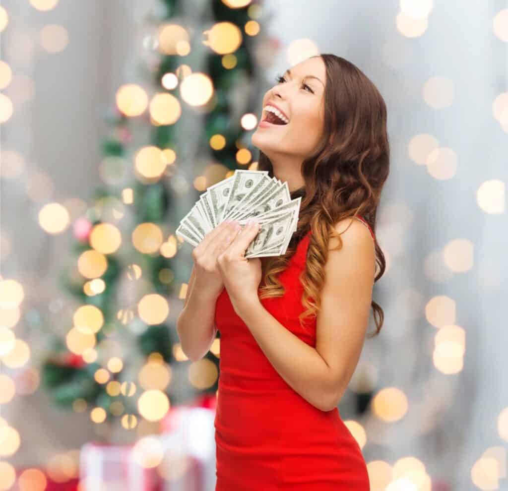 A woman in a red dress holds a fan of dollar bills, smiling with eyes closed, embodying the joy of stress-free Christmas savings. A blurred background of lights and a Christmas tree enhances the festive atmosphere.
