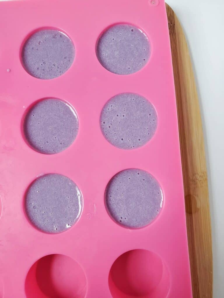 A pink silicone mold with six round cavities holds a purple sugar scrub mixture, ready to exfoliate, resting on a wooden board.