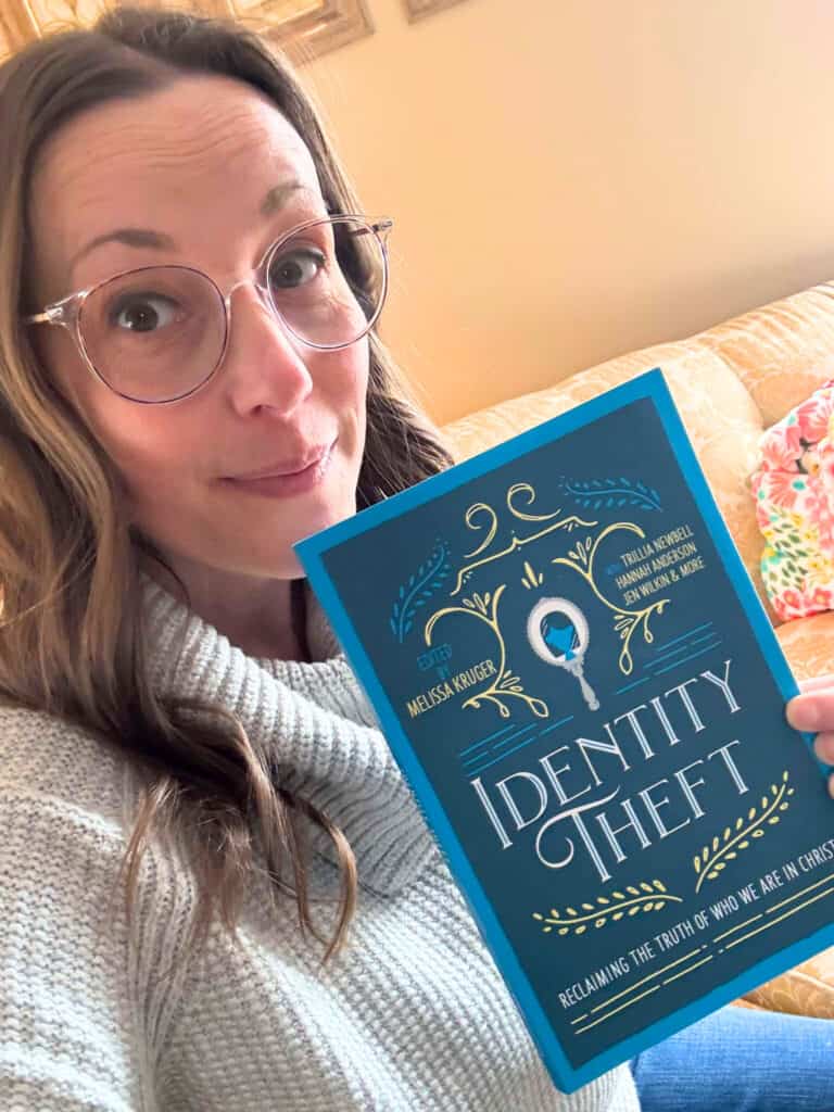 A person clad in a gray sweater and glasses sits indoors, engrossed in a book titled "Identity Theft," with a framed painting providing a touch of elegance. It's the perfect setting for unwinding after exploring the charming Bavarian Inn nearby.