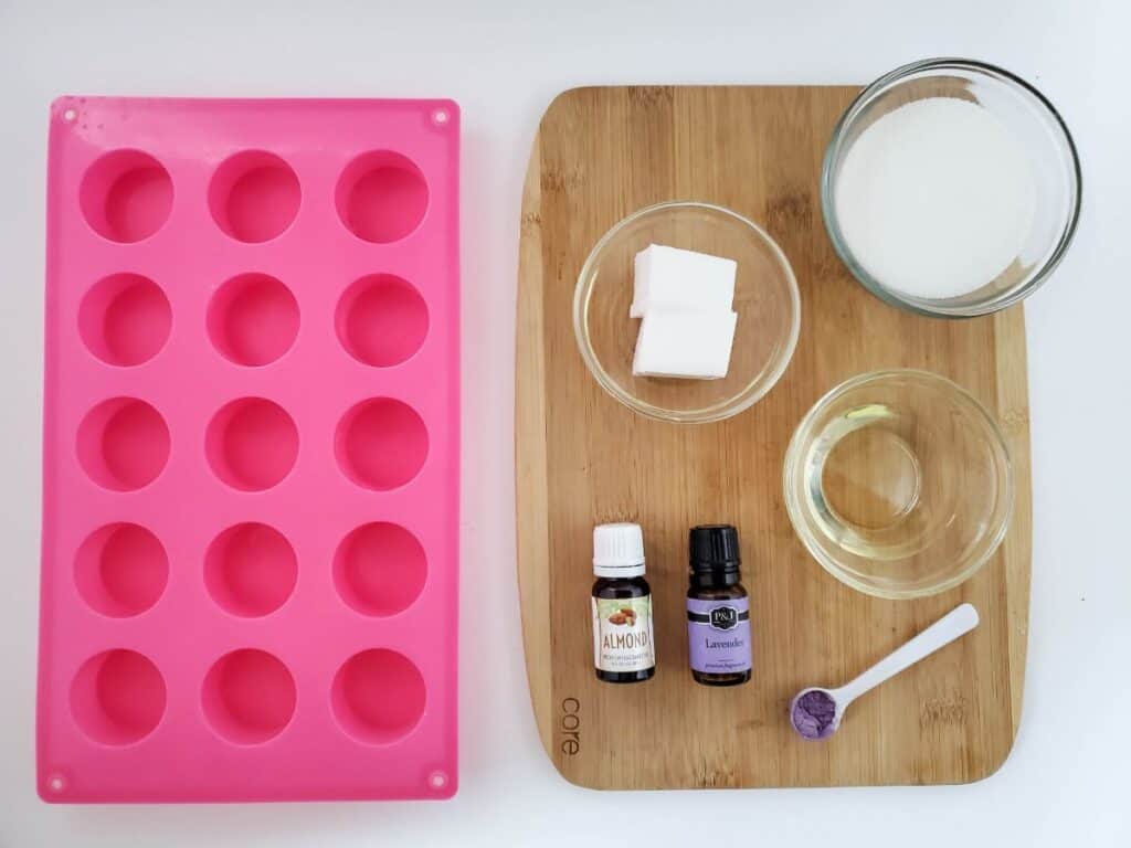 Silicone mold beside a wooden board with ingredients for exfoliating sugar scrub bars, including oil, lavender oil, almond extract, cubes, and purple powder in containers.