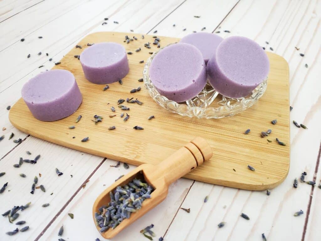 Four lavender exfoliating soap bars rest on a wooden board alongside a glass dish, with dried lavender artfully scattered around them.