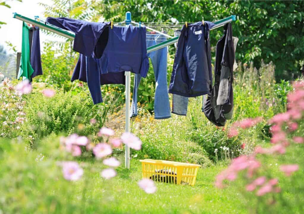 Clothes hanging on a rotary clothesline in a garden with a yellow basket on the grass, surrounded by green plants and pink flowers—a simple way to save money while doing laundry.