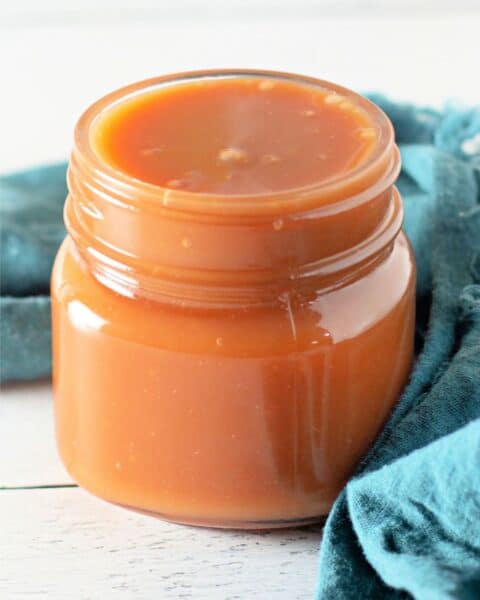 A jar filled with homemade caramel sauce is placed on a wooden surface next to a blue cloth, showcasing an easy caramel recipe.