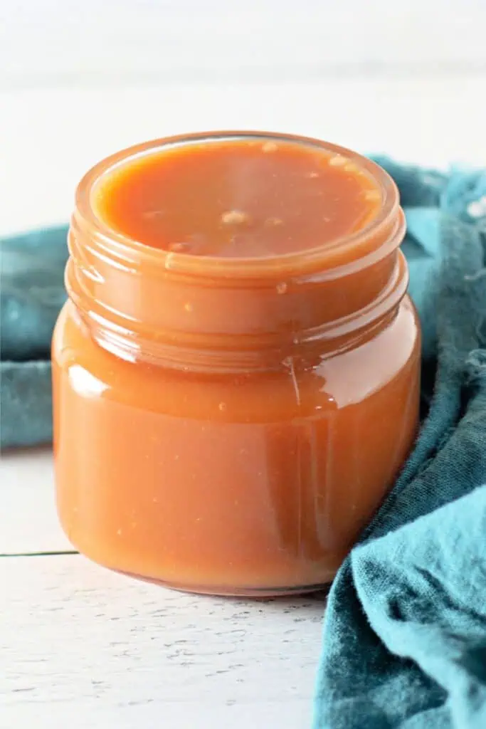 A jar filled with homemade caramel sauce is placed on a wooden surface next to a blue cloth, showcasing an easy caramel recipe.