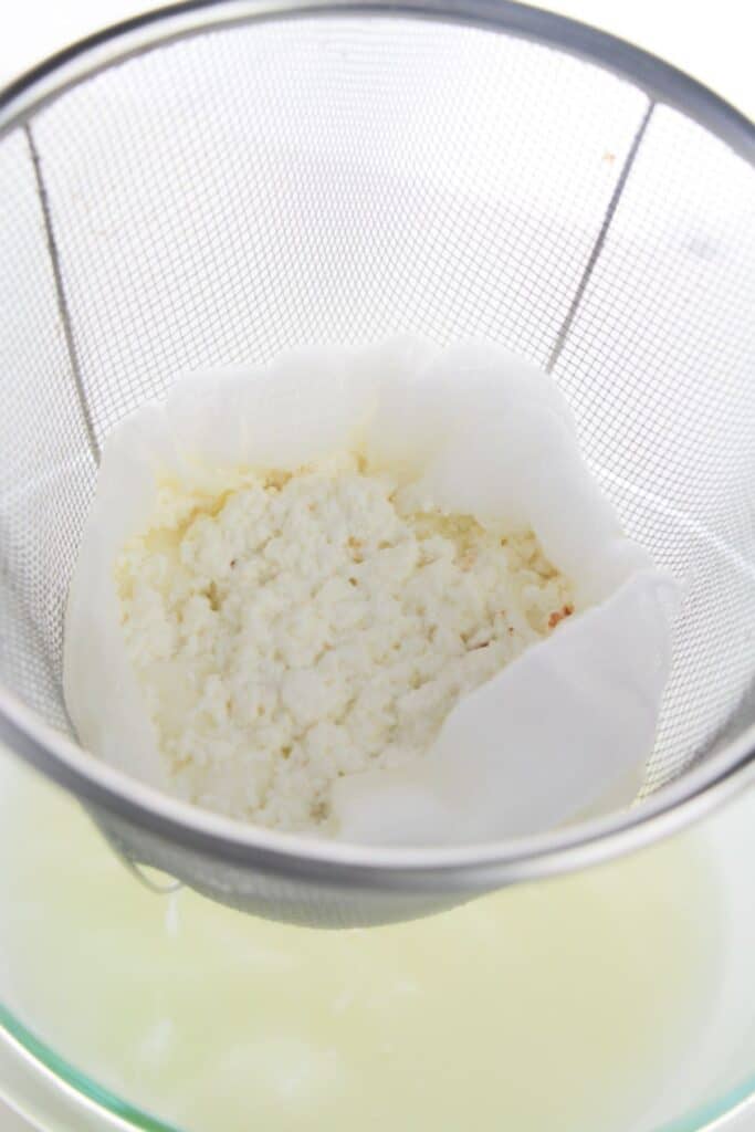 Curds are gently draining in a cheesecloth-lined strainer over a bowl, transforming the kitchen into a homemade haven as liquid whey separates from freshly made ricotta cheese.