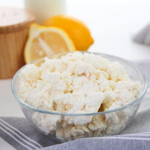 A glass bowl brimming with homemade ricotta cheese rests on a striped cloth. Behind it, a lemon wedge adds zest alongside a rustic wooden container, inviting you to explore this simple, delightful recipe.