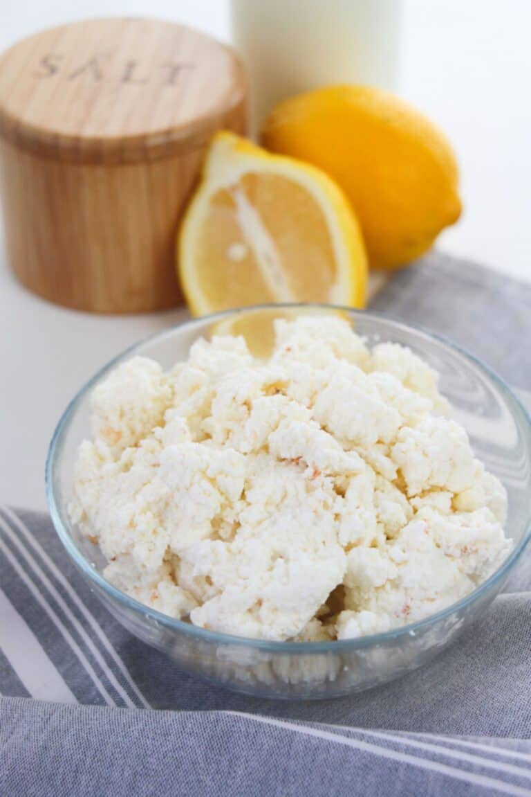 A bowl of homemade ricotta cheese sits on a striped cloth, accompanied by a halved lemon, a whole lemon, a wooden salt container, and a bottle in the background.