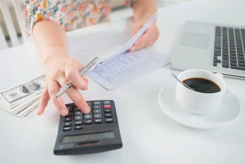 A person focused on financial planning uses a calculator while holding documents, surrounded by a laptop, cash, and a steaming cup of coffee on the table.