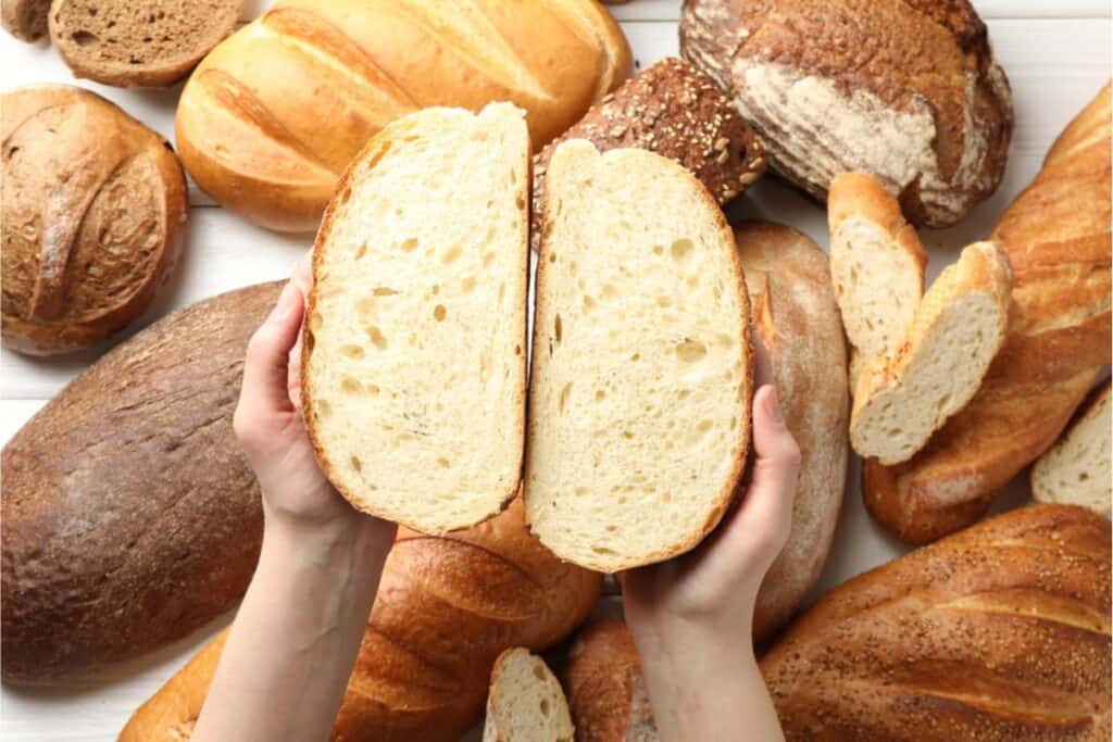Hands mastering two halves of a sliced sourdough loaf, surrounded by various whole loaves on a white surface.