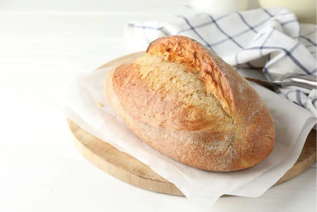 A loaf of freshly baked sourdough bread rests on parchment paper atop a wooden cutting board, with a blue and white checkered cloth in the background, capturing the essence of mastering sourdough baking.