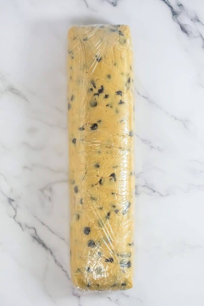 A rectangular block of chocolate chip cookie dough, reminiscent of a classic recipe, is wrapped in clear plastic on a marble countertop.