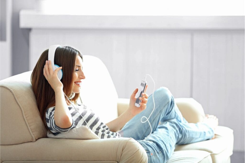 Woman in striped shirt and jeans enjoys cost-free home activities, listening to music with headphones while holding a smartphone, sitting on a beige couch with legs crossed.