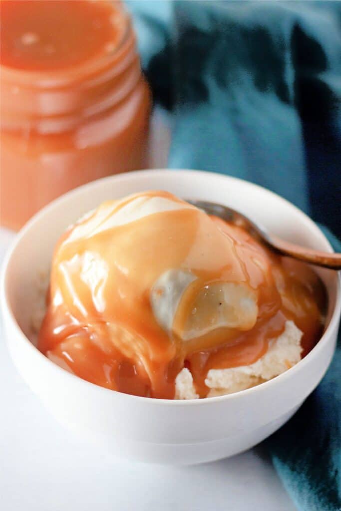 A bowl of dessert drizzled with a rich caramel sauce sits invitingly beside a jar brimming with an easy caramel recipe, accompanied by a gleaming spoon.