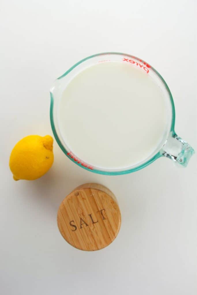 A measuring cup with milk, a lemon, and a round wooden container labeled "salt" sits on a white surface, the perfect setup for crafting homemade ricotta.