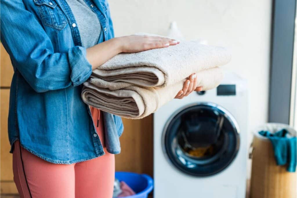 A person stands in the laundry room, expertly folding towels next to a washing machine and neatly organized baskets. Discover how to save money by mastering efficient laundry techniques that keep your clothes fresh without stretching your budget.
