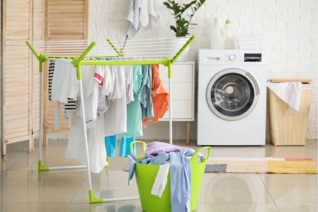 In the laundry room, clothes hang on a drying rack next to a green basket brimming with freshly washed items. The trusty washing machine sits nearby, alongside a laundry basket in the background—a simple way to save money by avoiding the dryer.