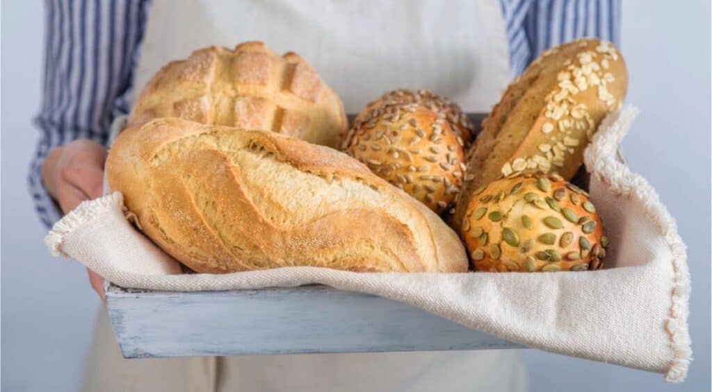 A person holds a tray with assorted breads, including loaves and rolls with seeds, on a cloth napkin—a perfect showcase for those following a beginner’s guide to mastering sourdough.